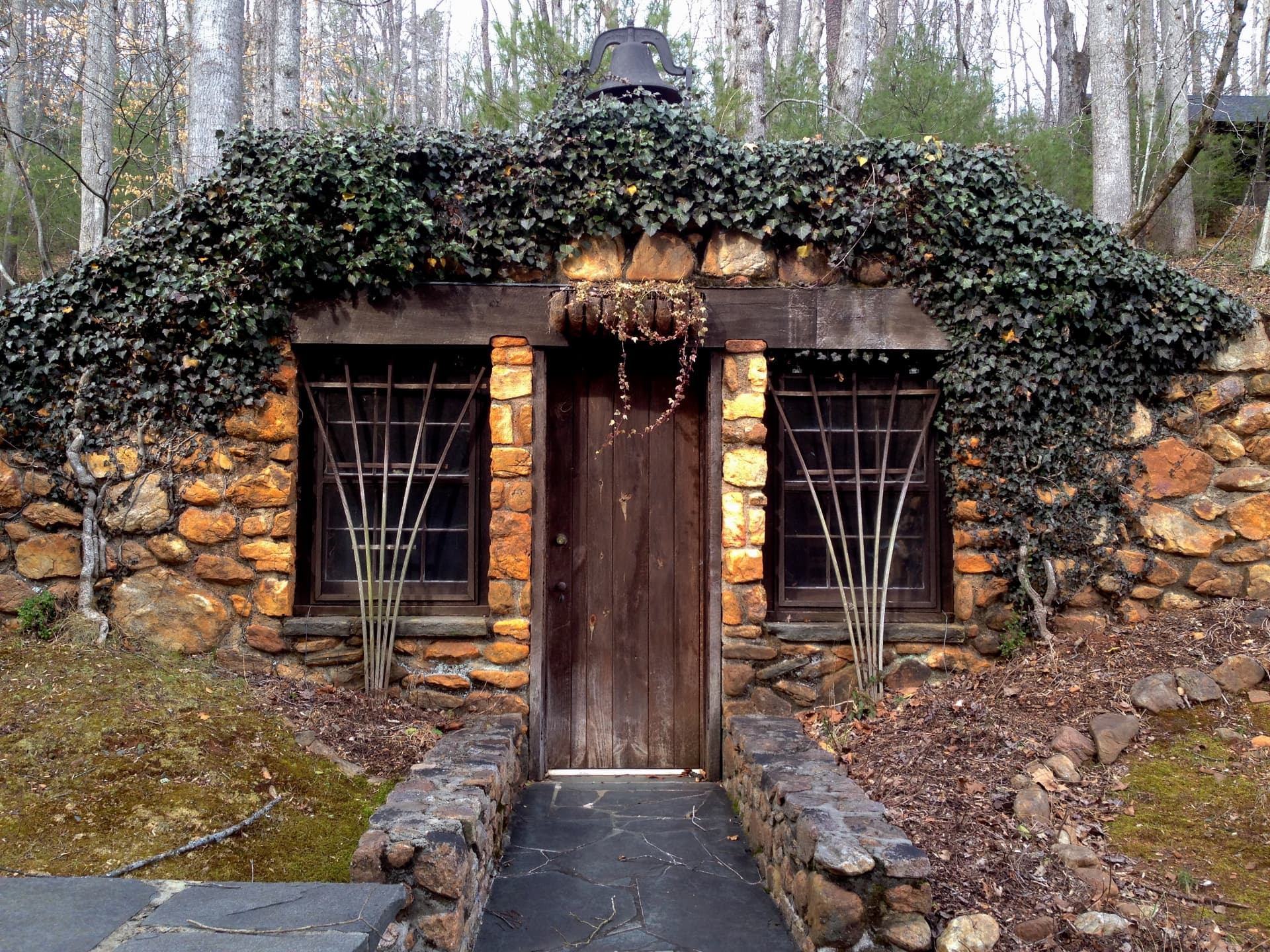 The ivy-covered stone gardener's storage house at Devotion, its low roof buried in ivy with a cast iron bell above the timber door and flagstone walk leading in