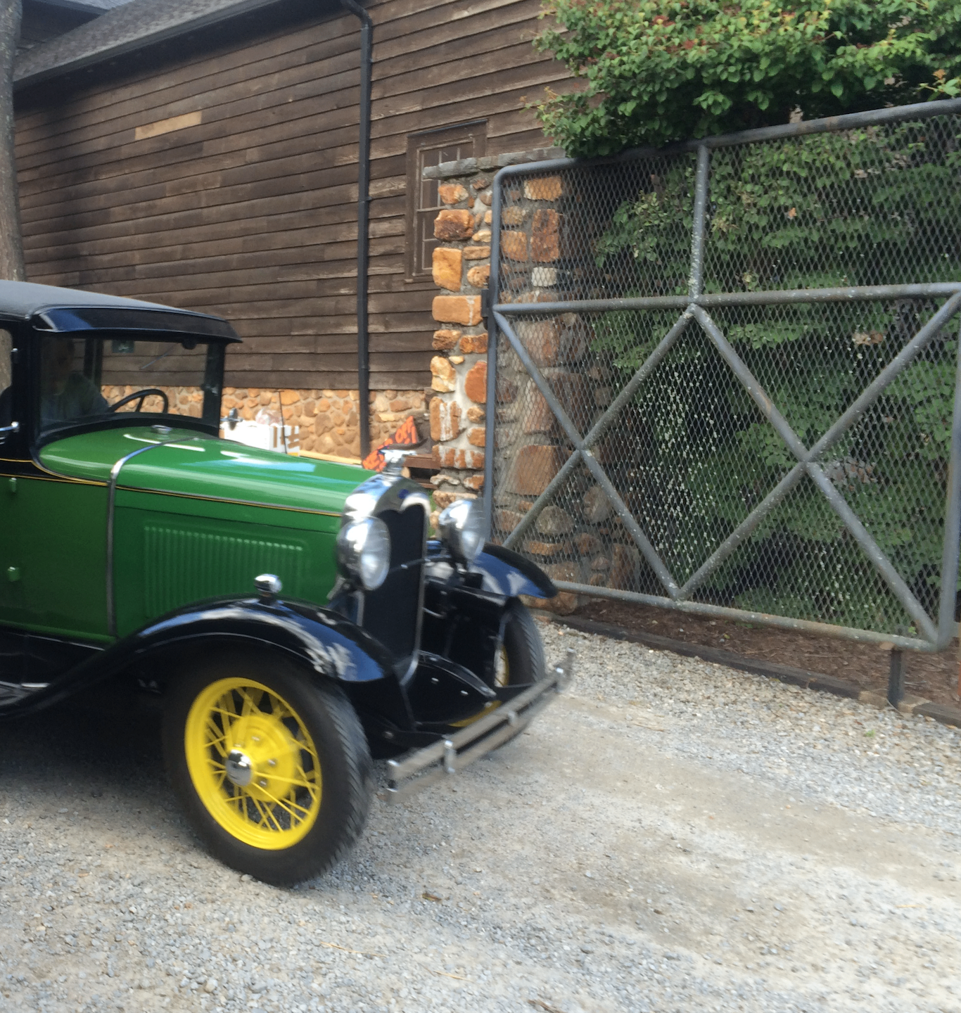 A green and black Ford Model A parked beside a chestnut-sided, stone-footed estate building
