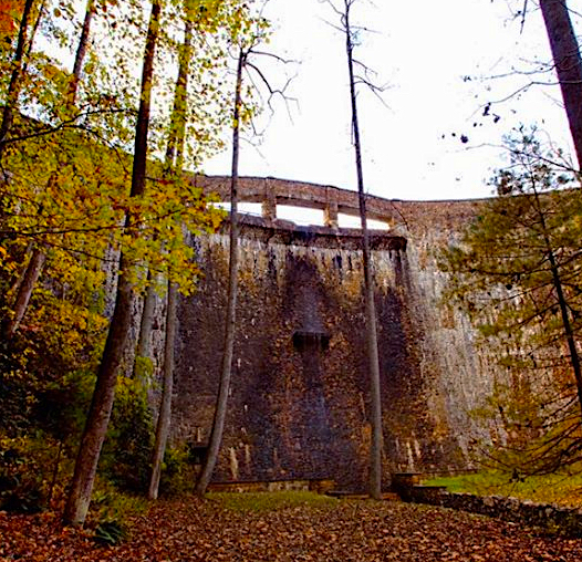 The downstream face of the Devotion hydroelectric dam rising through bare autumn trees, its stone wall crowned by the open parapet of the crest walkway