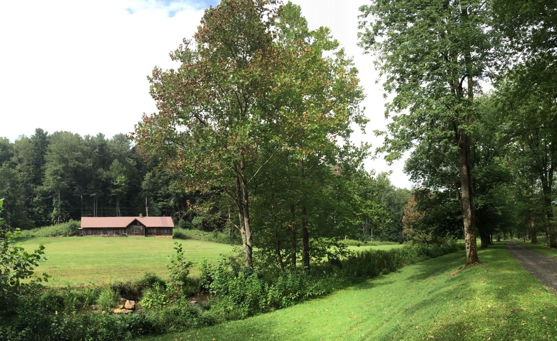 The long low poultry barn at Devotion with its red metal roof, seen across a wide mown field with hardwoods and a small creek in the foreground