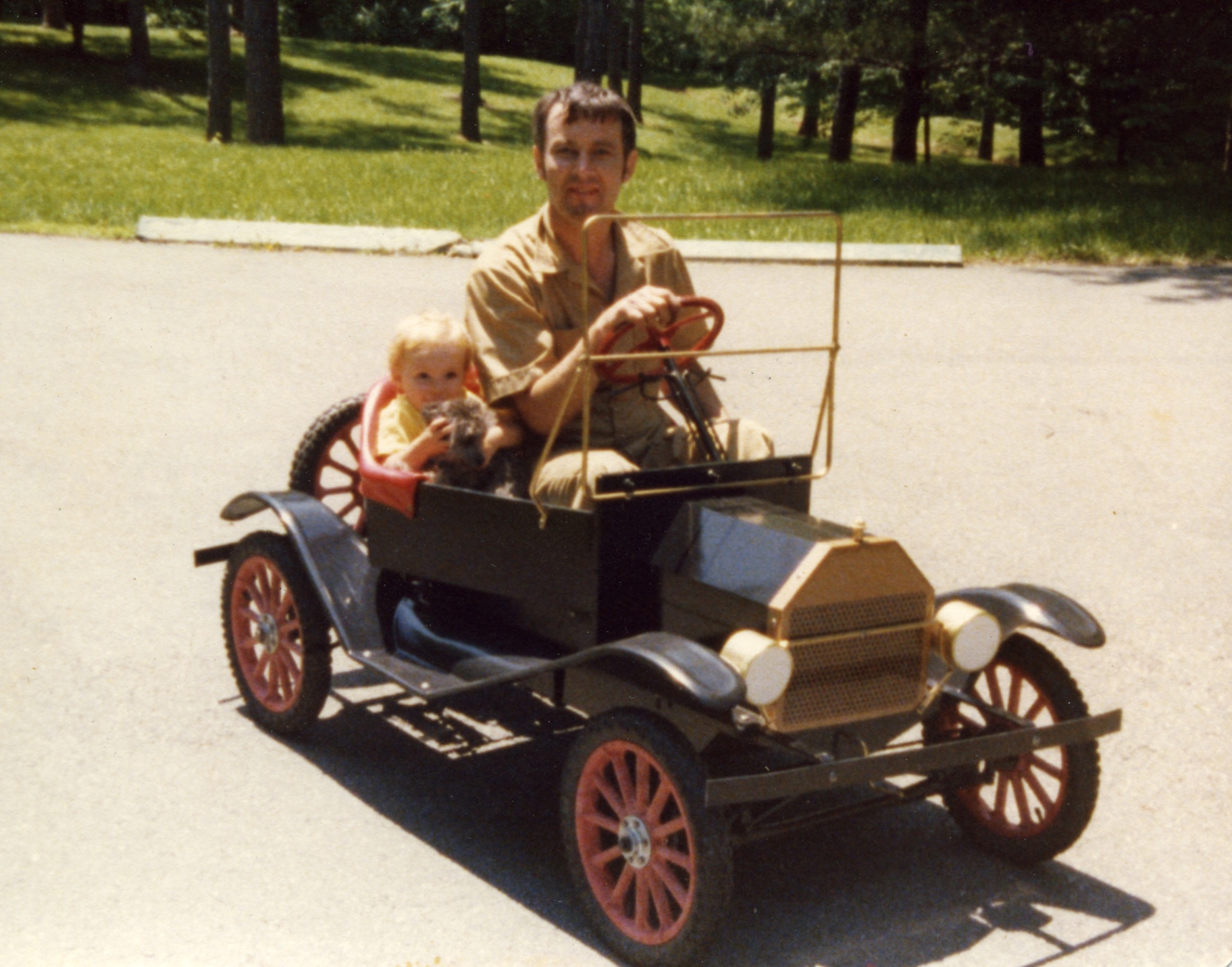 Zach Reynolds driving a small vintage roadster with his young daughter Kathryn and a dog beside him, 1978