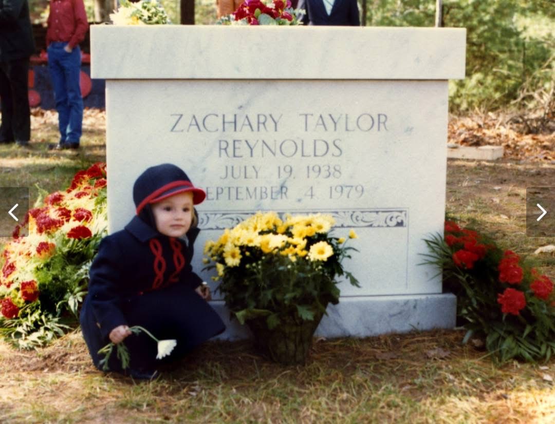 Young Kathryn Reynolds crouching beside her father's gravestone, inscribed Zachary Taylor Reynolds, July 19 1938 – September 4 1979