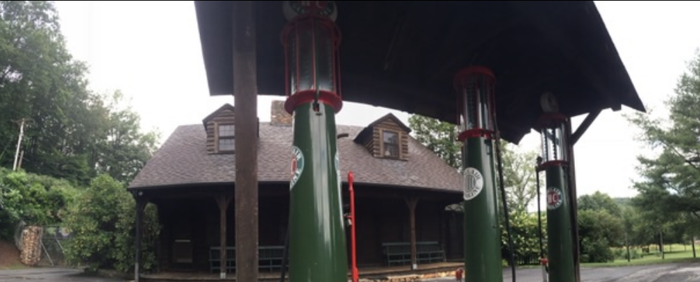 The log general store and post office at Devotion with a shingled roof, stone chimney, vintage Sinclair gasoline pumps, and a black 1930s sedan beneath the shed roof