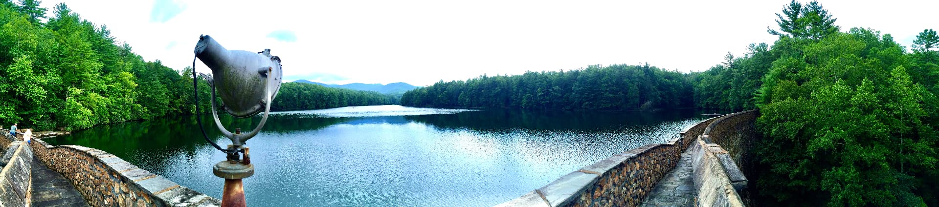 Panorama from the stone walkway atop the Devotion dam looking up the lake, its still water ringed by forested ridgelines, with an old floodlight mounted on the parapet