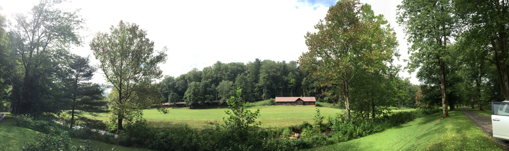 Wide panorama of the open bottomland at Devotion where the chicken houses stood, a long chestnut barn and outbuildings at the far edge of a mown field ringed by hardwood forest