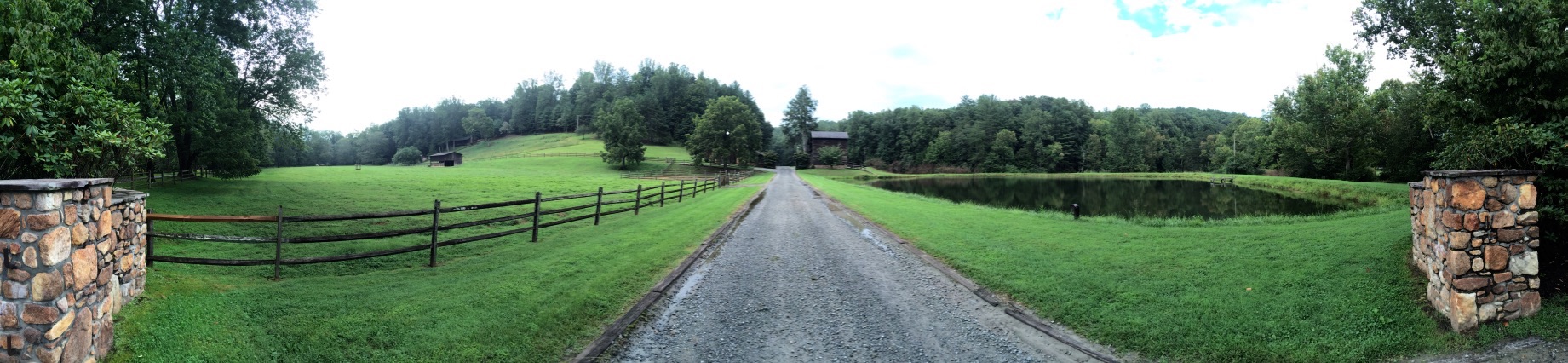 Panorama looking down the gravel road to the barn at Devotion, between stone gate pillars, with fenced pasture and a pond on either side and the barn standing among trees at the far end