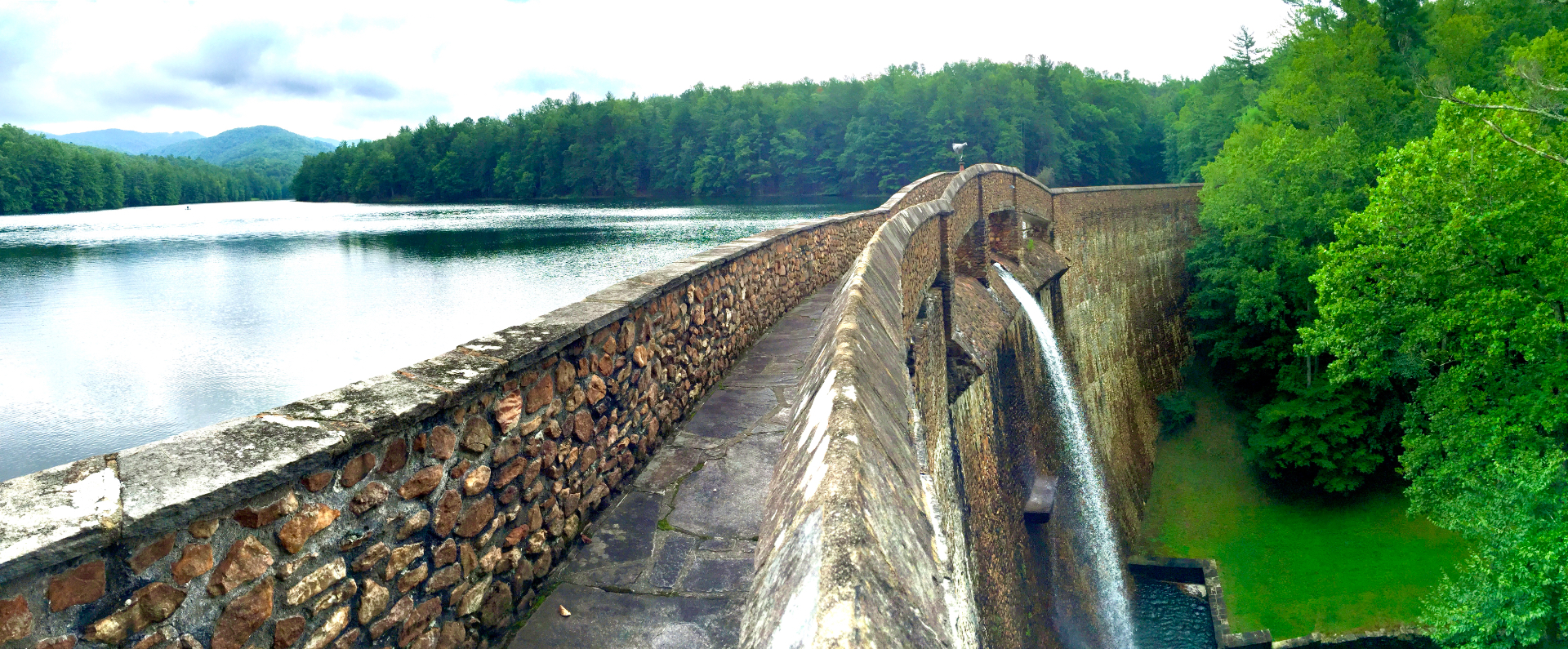Stone walkway along the crest of the Devotion hydroelectric dam, with the mile-long lake and foothill ridgelines to the left and the spillway falling into the gorge to the right